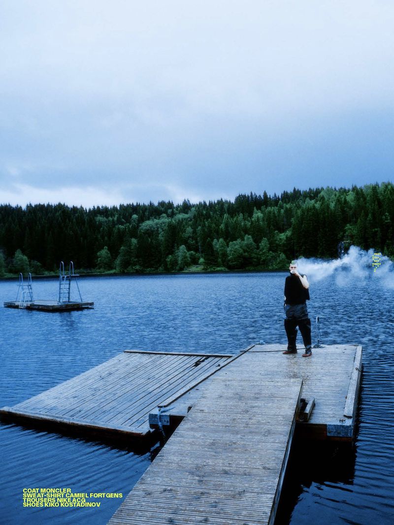 A man stands on a wooden dock at a lake's edge on an overcast day, with a forest and cloudy sky in the background, fashion shot.