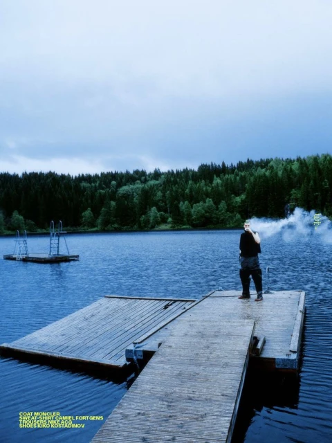 A man stands on a wooden dock at a lake's edge on an overcast day, with a forest and cloudy sky in the background, fashion shot.