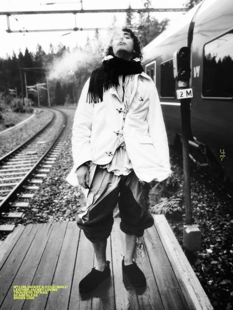 Monochrome portrait of a slender man standing on a platform, smoking with a train behind him. He wears a jacket, scarf, and shoes.