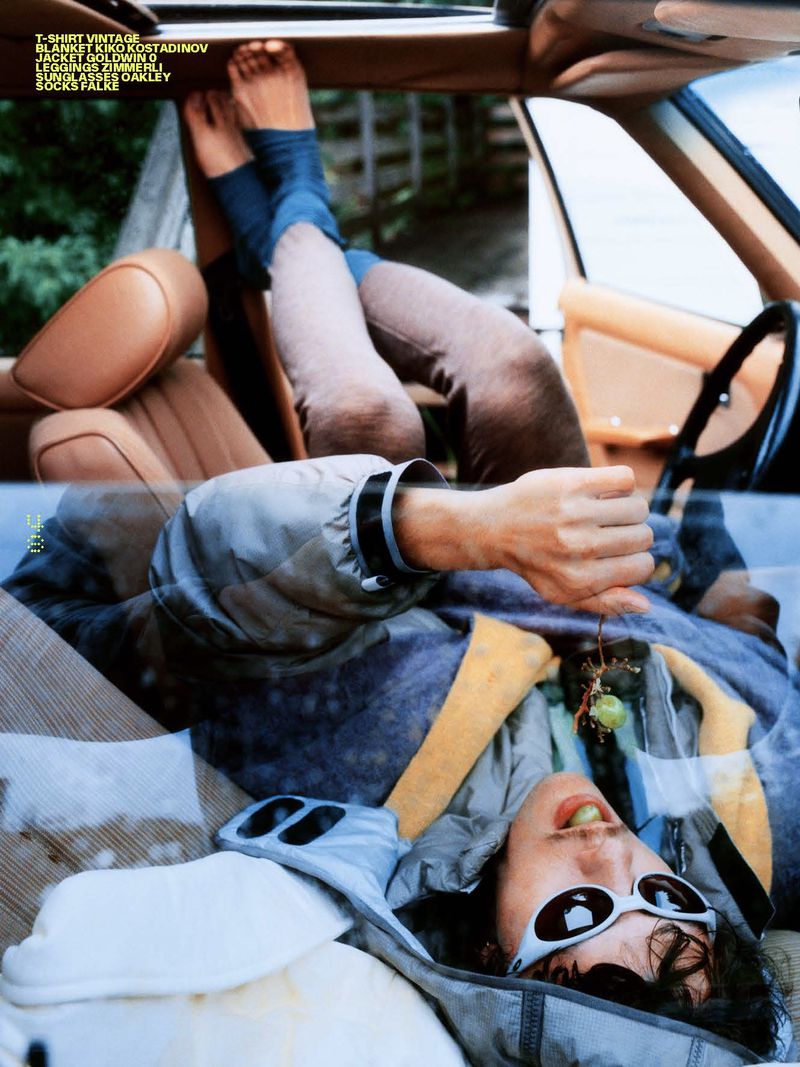 Man in white sunglasses and a hooded jacket lying across car seats with bare feet elevated while eating a green grape.