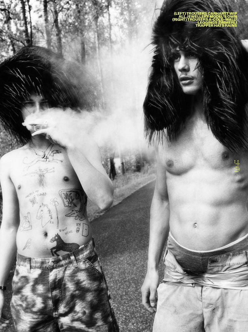 Black and white fashion shot of two shirtless men with fuzzy hats standing outdoors, one blowing smoke in a forest setting.