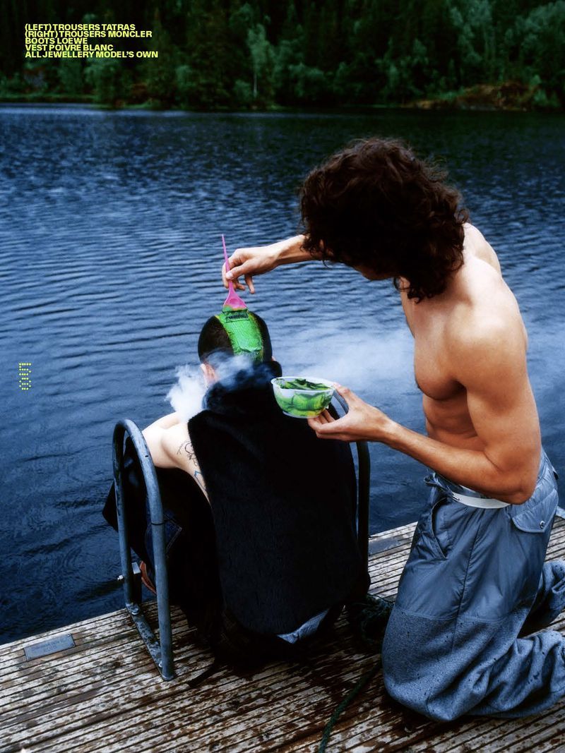 Two men on a wooden dock by a lake: one shirtless pouring from a bowl on the other man's head, dark trees visible in the background.