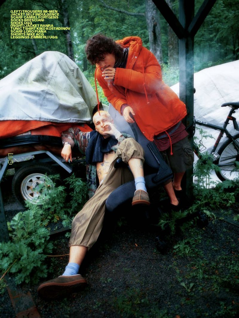 Two men in an outdoor setting. One man is lying down and smoking while another man stands beside him in an orange jacket.