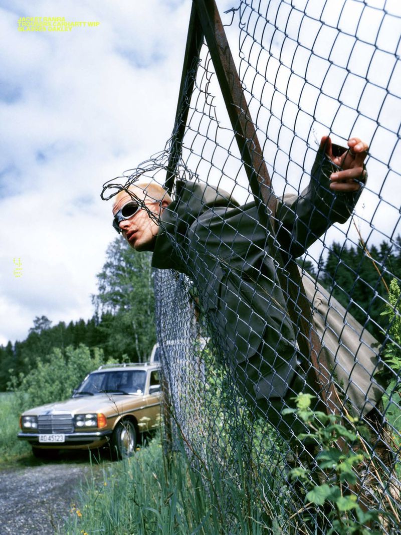 A bald adult man in sunglasses and a hooded jacket climbs through a chain-link fence, with a gold car and trees visible in the background.