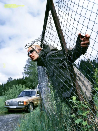 Model wearing wrap sunglasses and a hooded olive jacket leaning through a chain-link wire fence near a parked vintage car.