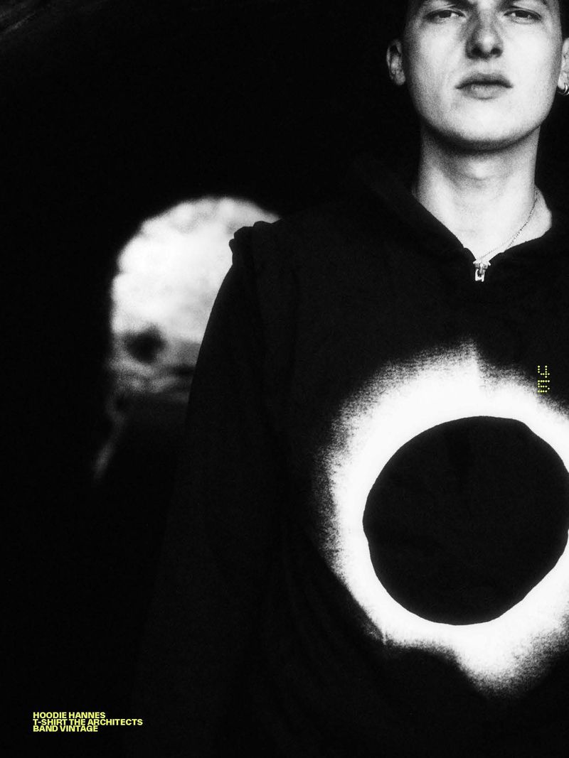 Close-up portrait of a young man wearing a black hoodie with a white spray-painted circle graphic against a dark background.