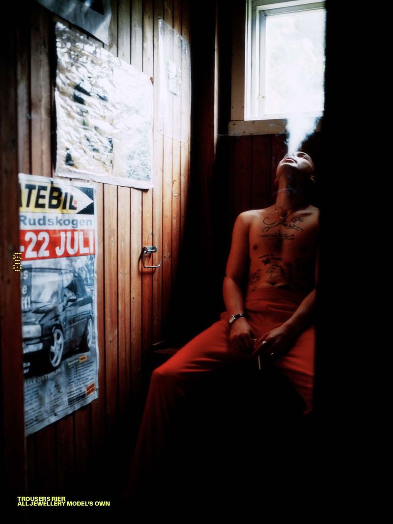 Shirtless man with chest tattoos in red trousers exhales smoke inside a wood-paneled room beside a window.