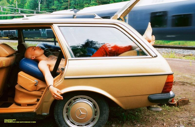Adult male with bare chest lounges in tan station wagon with luggage; legs stretch into open hatchback near passing train and green forest.