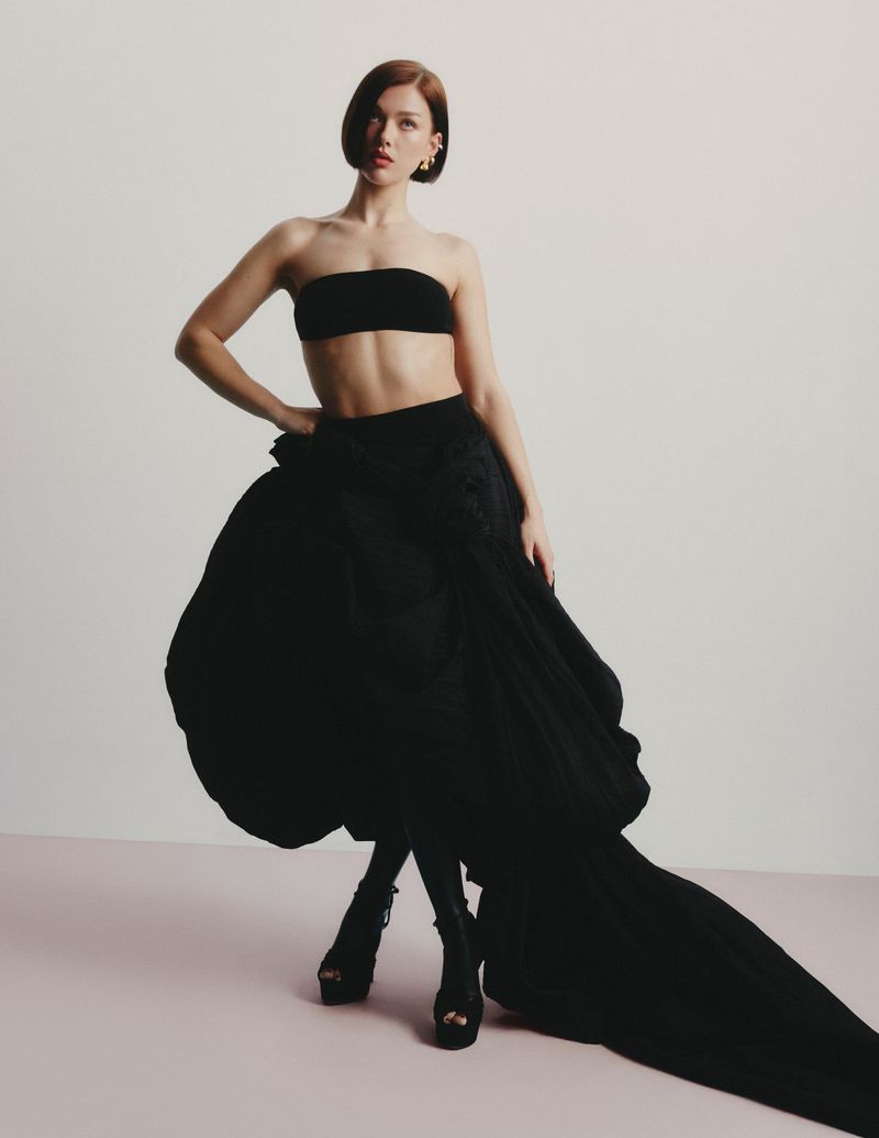 A woman poses in black bandeau top and flowing skirt with platform heels against a gradient pink and white backdrop in studio lighting.