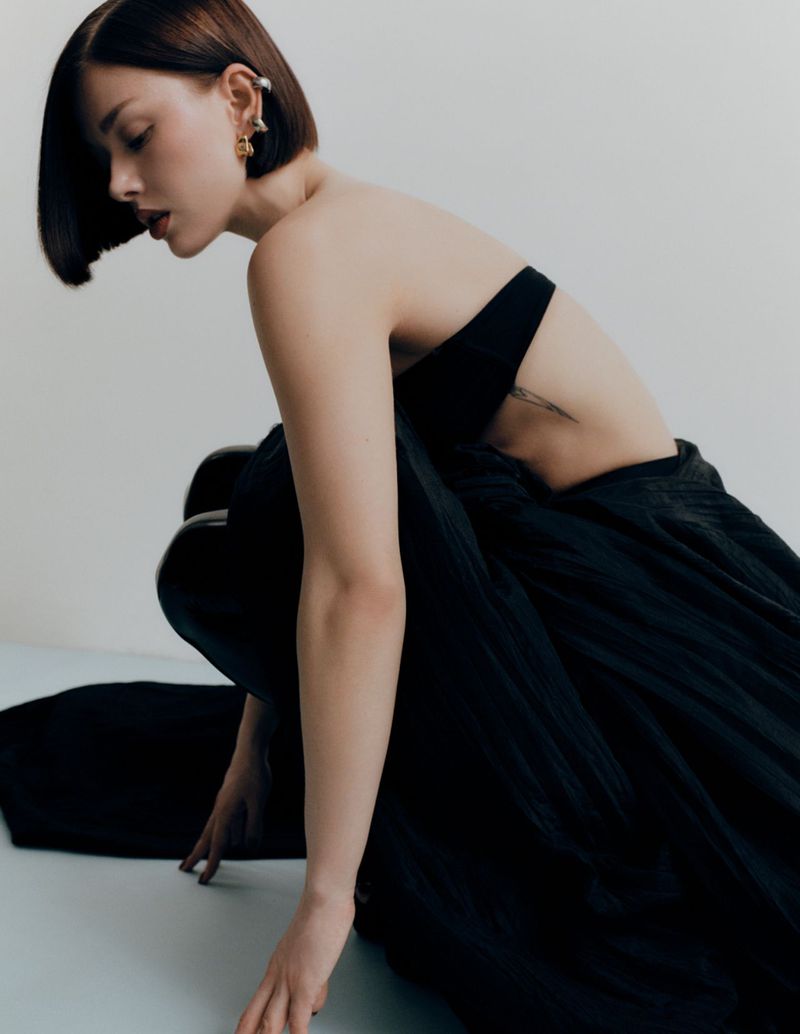 Portrait of a young woman with dark hair posing in a strapless black dress against a white background, studio lighting, fashion editorial.