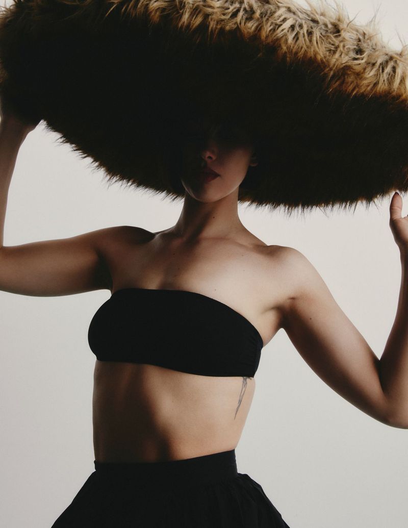 Model with oversized furry hat and black two-piece outfit poses in a studio, showcasing fashion and avant-garde style on a light background.