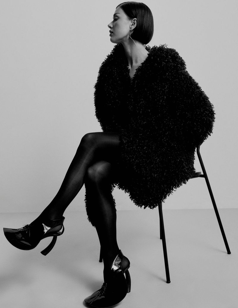 Monochrome fashion shot of a woman with short dark hair in a fur coat and patent heels, sitting on a black chair in a photo studio.