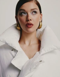 Jess Alexander wearing a voluminous white high-collar puffer coat and chunky gold dome earrings against a neutral studio backdrop.