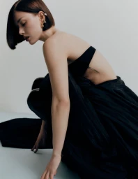 Jess Alexander crouching in a black strapless gown with a sleek dark bob haircut against a minimalist studio background.