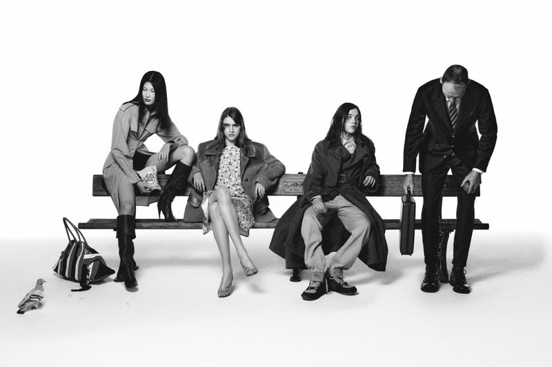 Four fashionably dressed adult women and a man pose on a park bench in a black and white fashion editorial, with a pigeon at their feet.