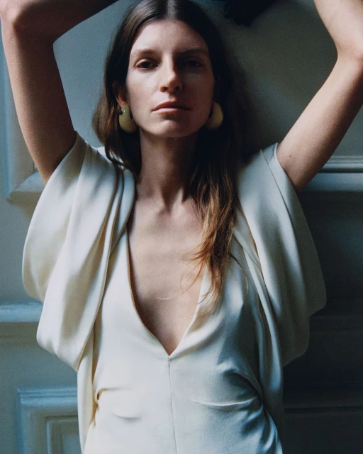 Close-up of a stylish, adult woman with long brown hair posing indoors, wearing a light-colored dress and hoop earrings in soft light.