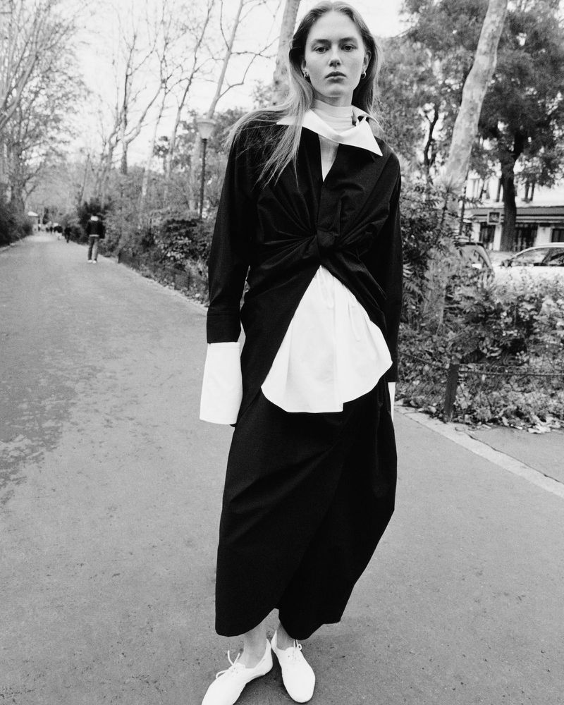 Black-and-white outdoor fashion shot of a slender woman in a black and white outfit standing on a park path surrounded by trees.
