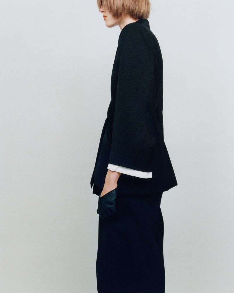 Side profile studio shot of person with blond hair wearing a black skirt suit with gloves against a plain white backdrop.