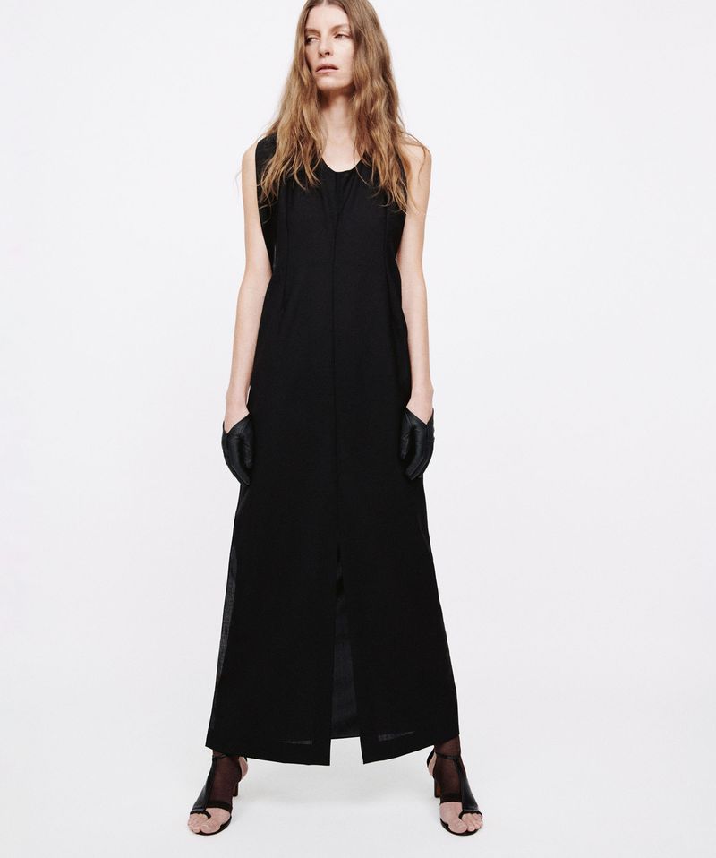 Full-length studio shot of a woman posing in a black dress, gloves, and sandal heels against a white background. Fashion editorial.