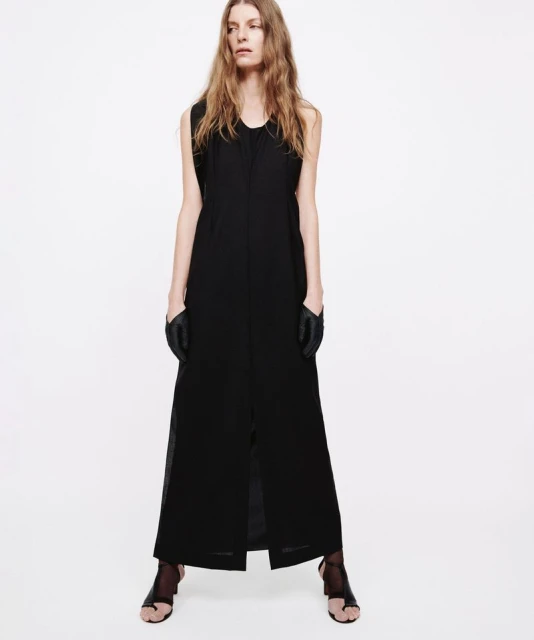 Full-length studio shot of a woman posing in a black dress, gloves, and sandal heels against a white background. Fashion editorial.