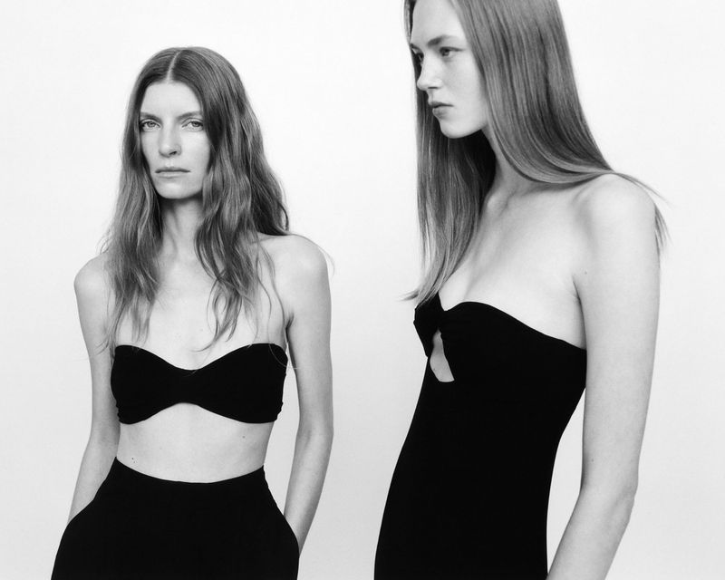 Two women pose in black strapless dresses against a white backdrop, showcasing minimalist fashion and classic style in a studio setting.