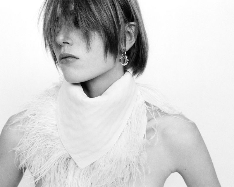 Black-and-white studio portrait of a person with short hair wearing feathered scarf, gazing off-camera, minimalist aesthetic.