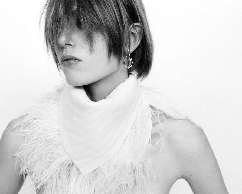 Black-and-white studio portrait of a person with short hair wearing feathered scarf, gazing off-camera, minimalist aesthetic.