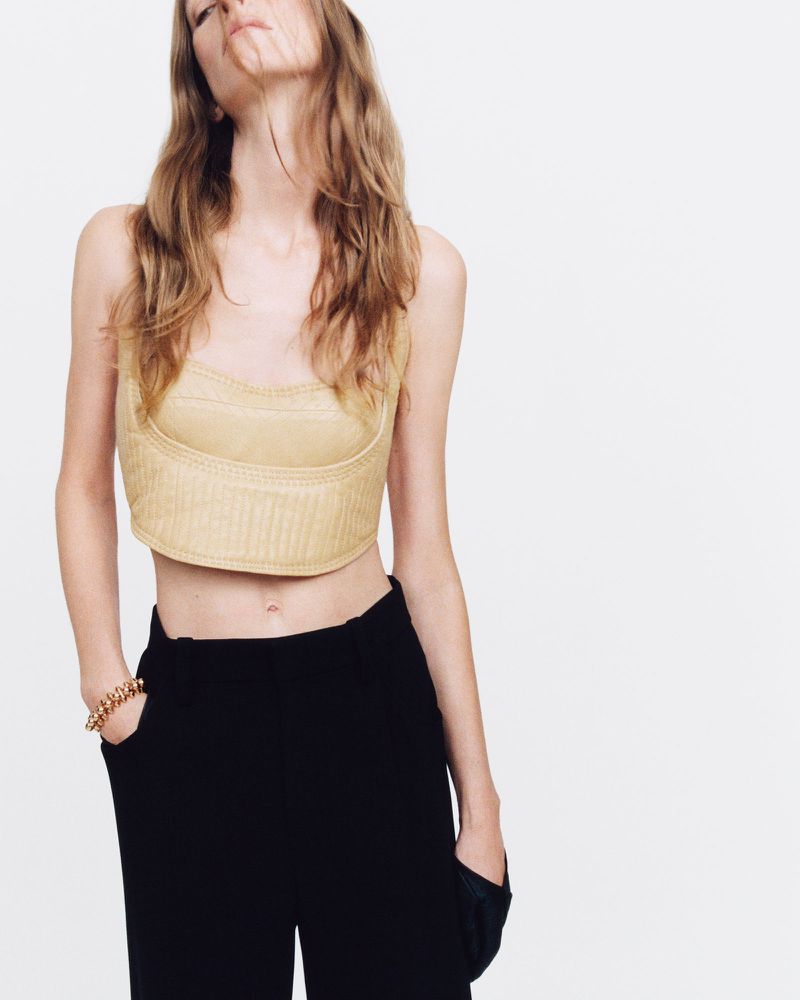 Fashion shot of a fair-skinned adult female wearing a yellow corset top and black slacks in a studio with a white background.