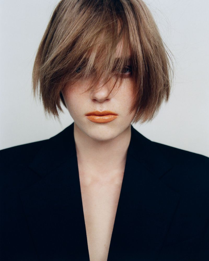 Close-up shot of a young adult woman with blonde hair and black blazer against a white background. Fashion, beauty, and portrait photography.