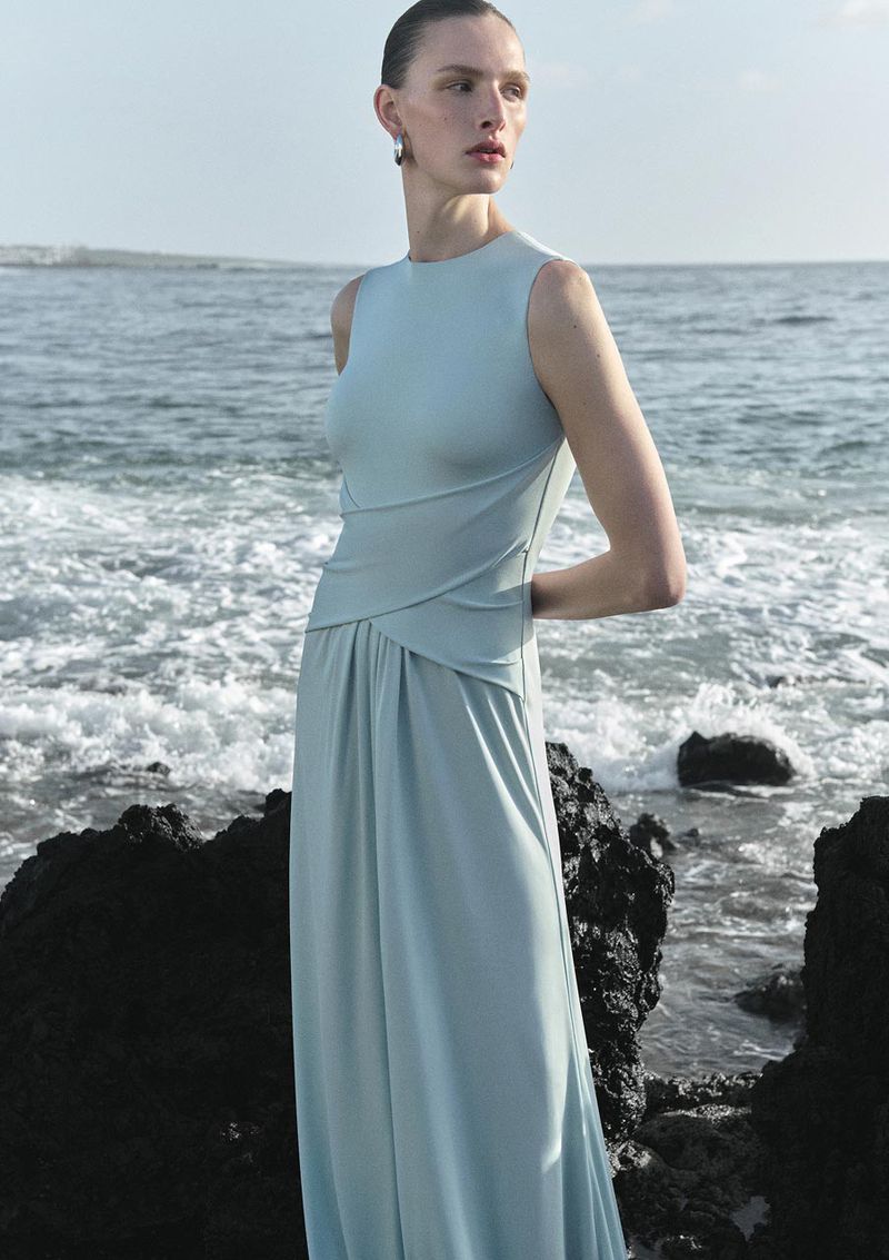 Fashion shot of woman in light blue gown posing on rocky beach near ocean with natural lighting and soft color palette for editorial mood.