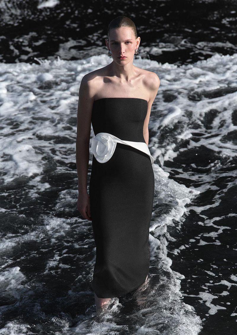 Woman in black strapless dress with white detail poses in water at the edge of the sea, accessorized with earrings.