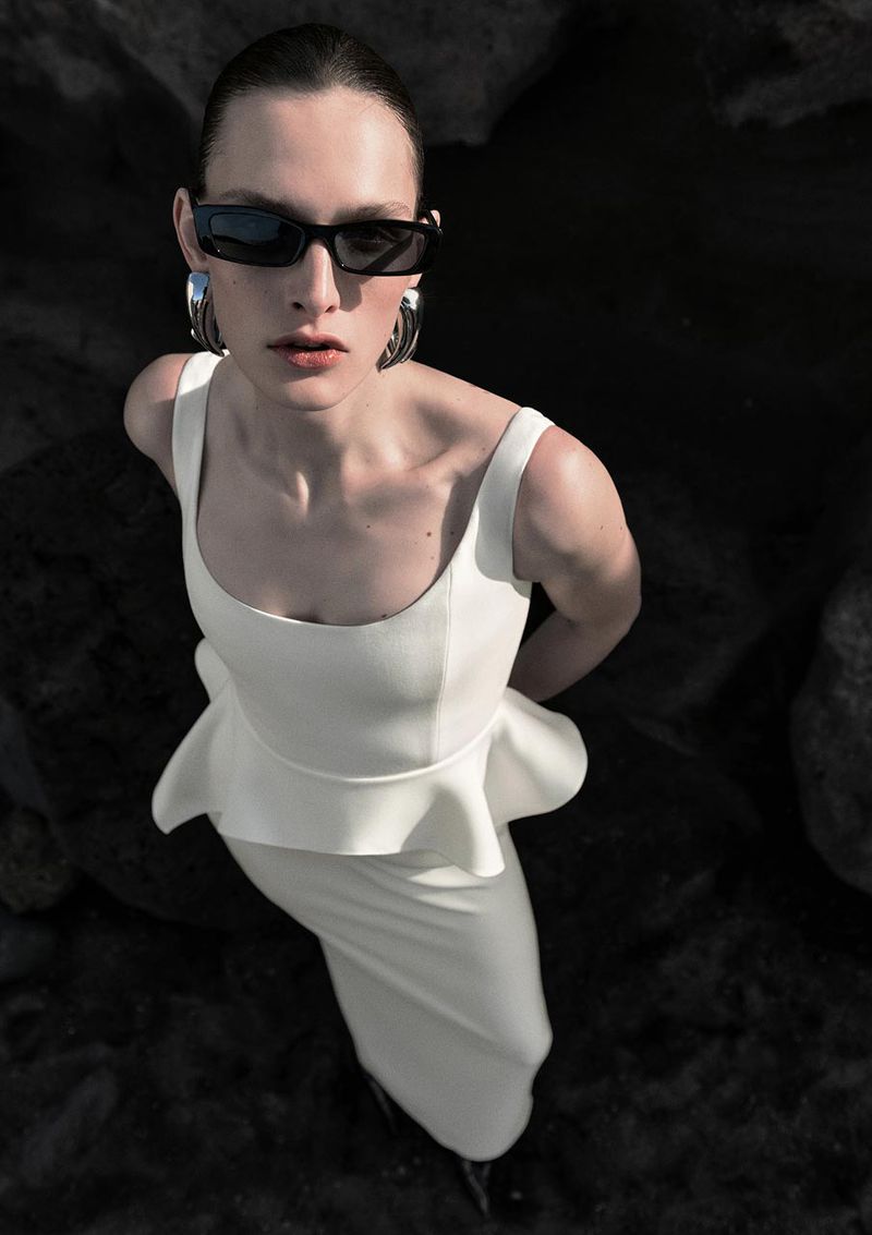 Woman in white dress, sunglasses, and silver earrings stands against a dark rocky background in an eye-level fashion shot.