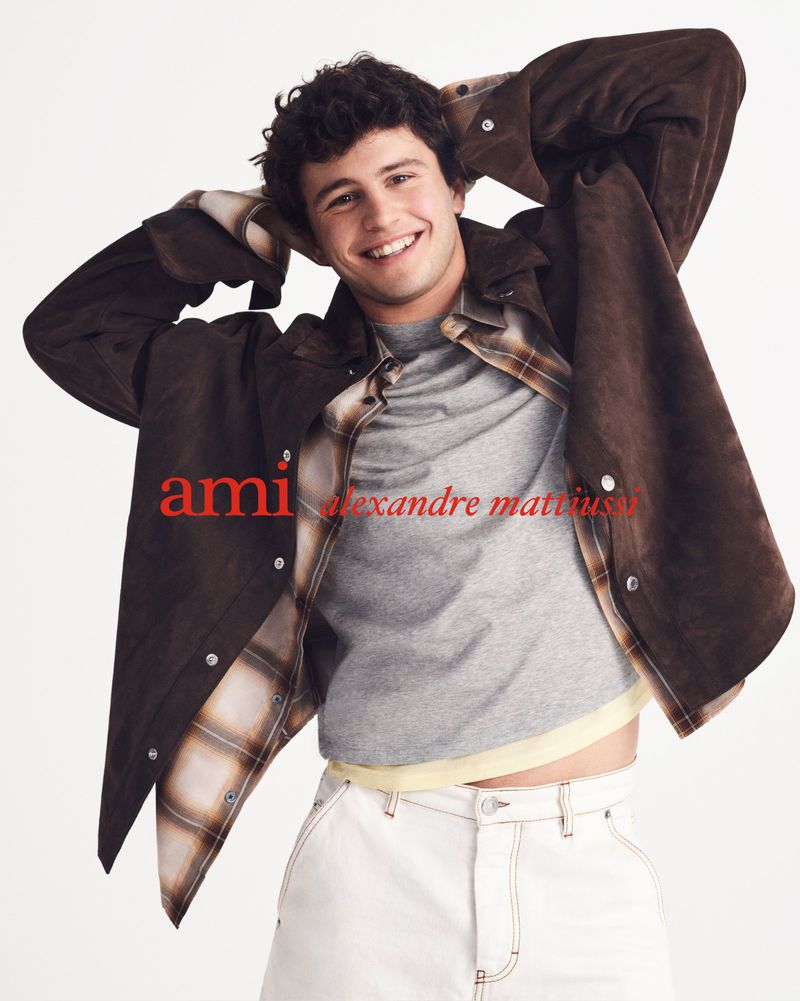 A smiling young man in a brown jacket and white pants poses in a studio with white background. The logo 'ami' is visible.