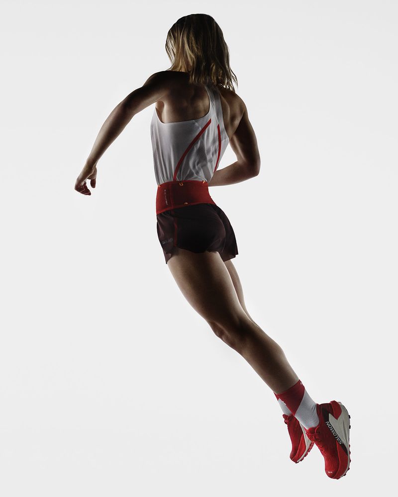 Female athlete in motion, viewed from behind. She wears a tank top, shorts, red belt, red shoes, and socks, against a bright, neutral background.