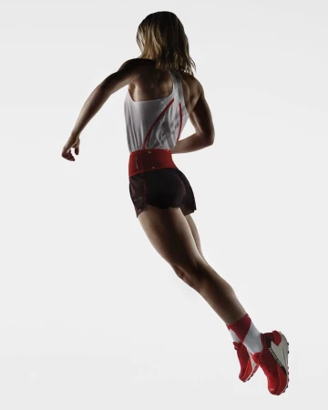Female athlete in motion, viewed from behind. She wears a tank top, shorts, red belt, red shoes, and socks, against a bright, neutral background.