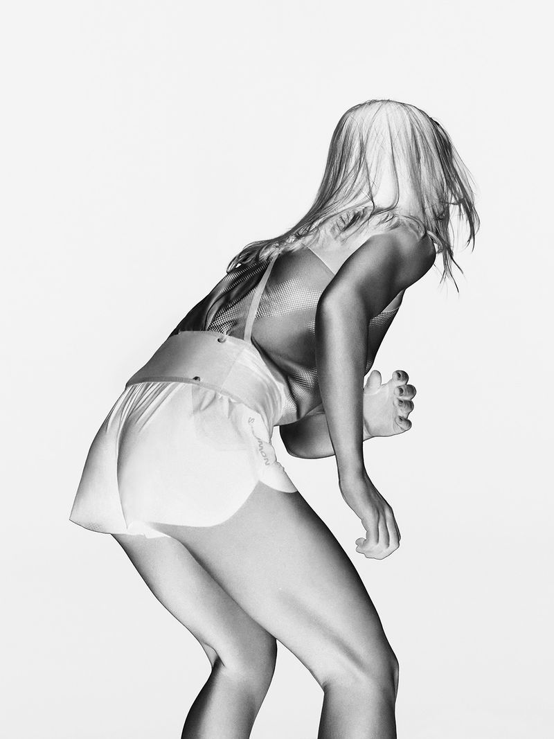 Monochrome shot of a woman with blond hair, wearing a sports bra and shorts, leaning forward with one arm raised and fingers bent.