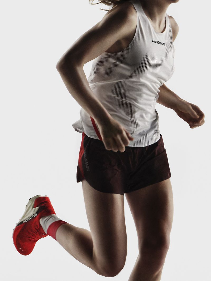 Athletic woman in motion, wearing a white tank top and maroon shorts, running shoes, in a studio setting against a clean white backdrop.