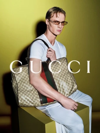 An adult male wearing a white shirt and sunglasses is sitting on a yellow box holding a Gucci bag against a yellow background.
