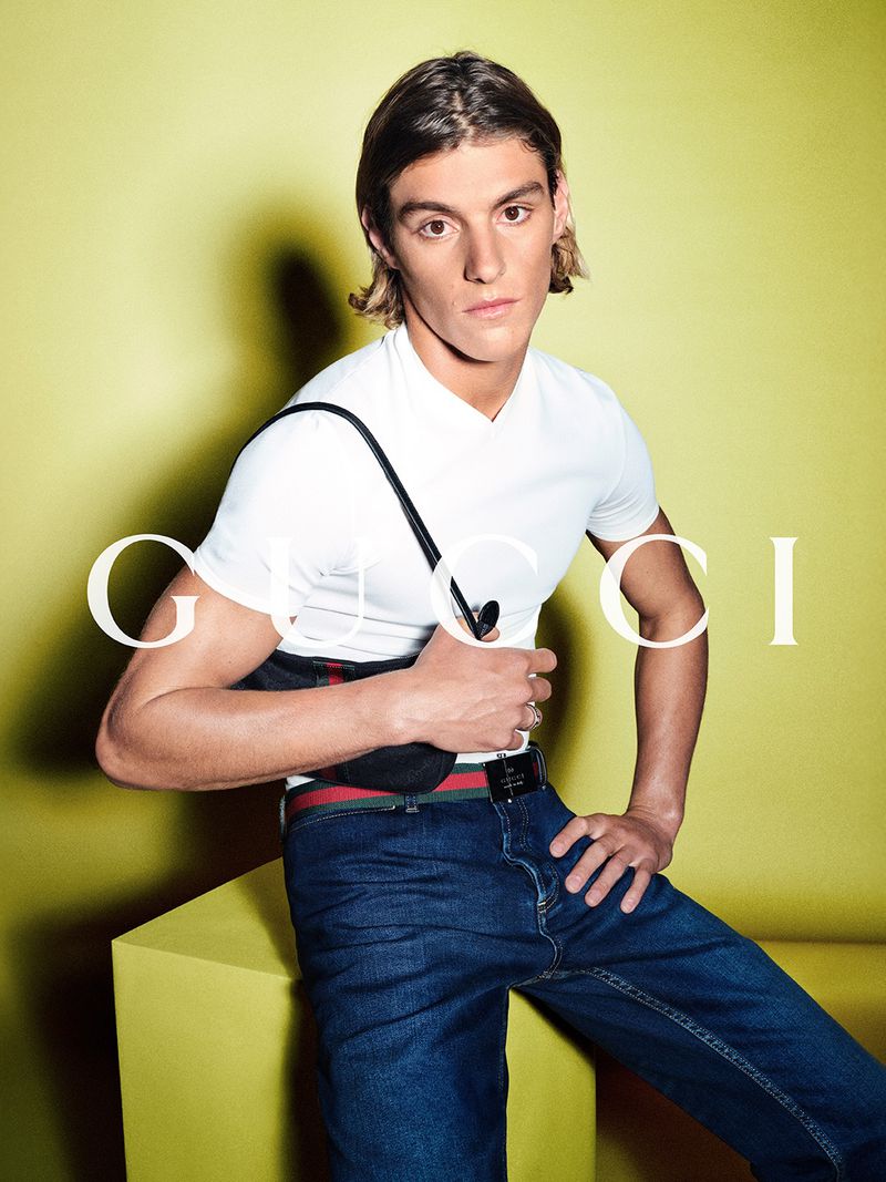 A brunette adult male poses in a studio with a yellow backdrop, wearing a white V-neck shirt, jeans, and a black crossbody bag.