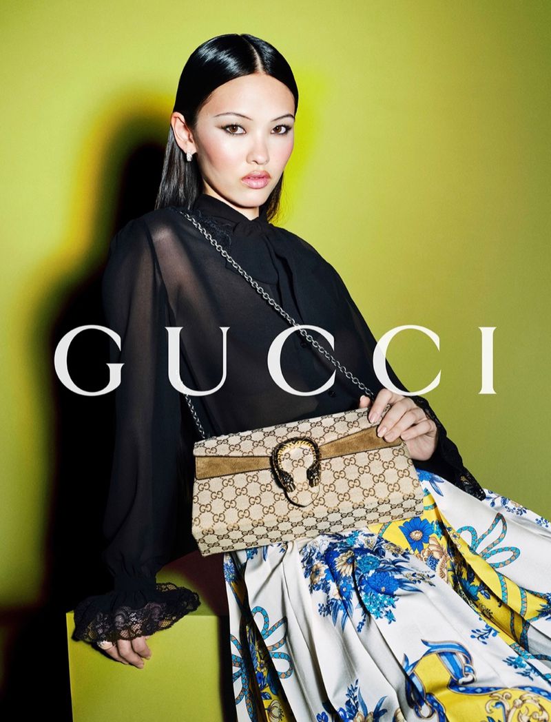 A woman in a black top and floral skirt poses with a Gucci handbag against a lime green backdrop in a fashion studio shot.