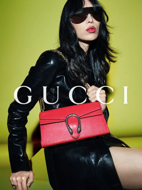 Fashion shot of a woman in a black outfit with a red purse and sunglasses, set against a bright yellow background.