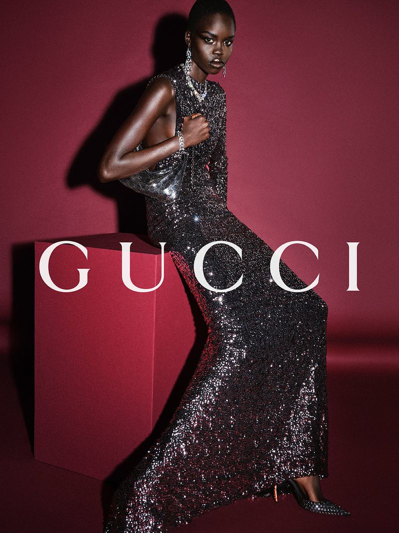 A striking dark-skinned model in a glittering gown poses against a deep red background, holding a metallic purse, and showcasing jewelry.