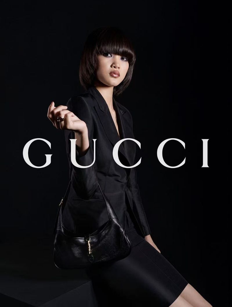 A young Asian woman poses in a black suit with a Gucci purse in a studio fashion shot, set against a dark background with Gucci logo.