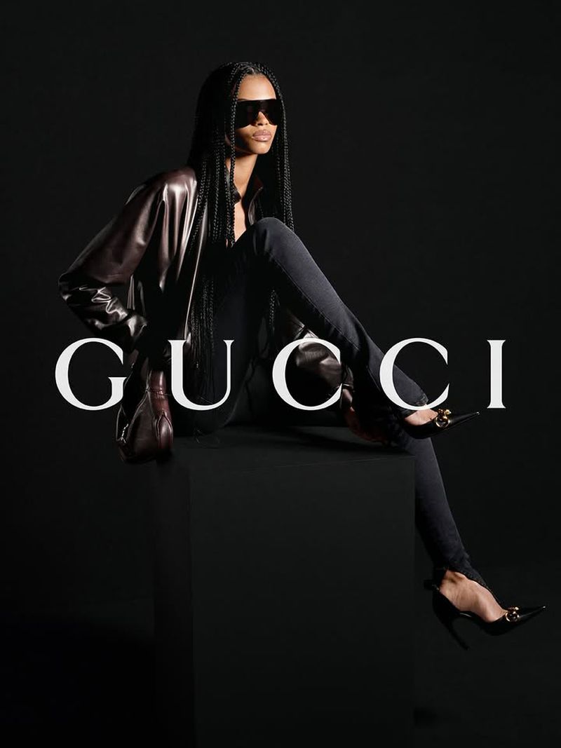 Fashion shot of a Black female model in sunglasses and a leather jacket sitting in a studio with a dark background and the Gucci logo.