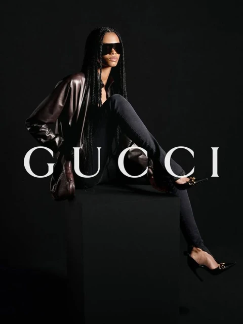 Fashion shot of a Black female model in sunglasses and a leather jacket sitting in a studio with a dark background and the Gucci logo.