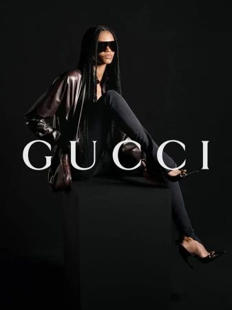 Fashion shot of a Black female model in sunglasses and a leather jacket sitting in a studio with a dark background and the Gucci logo.