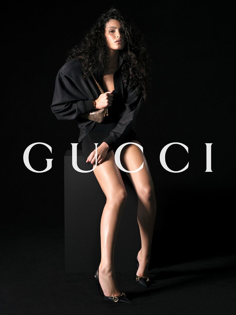 Fashionable woman poses in a black dress and jacket, seated against a black backdrop. She carries a small gold handbag and wears black heels.