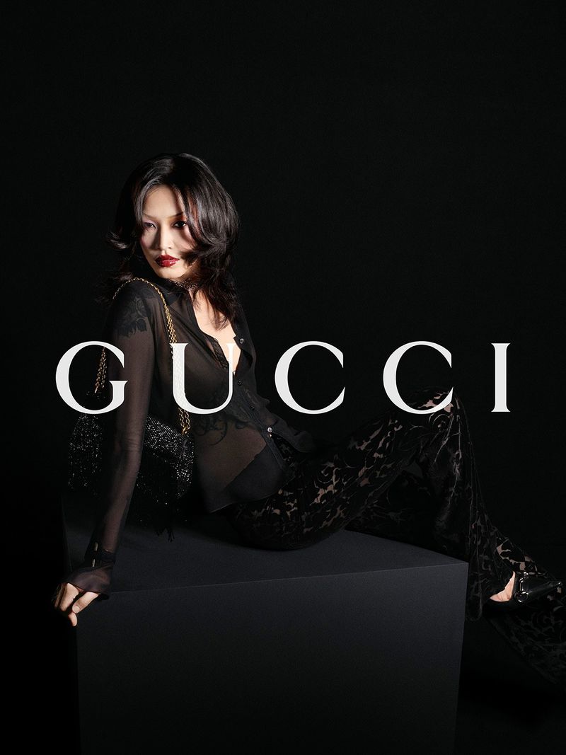Asian woman poses in a semi-sheer black blouse and patterned trousers, seated against a dark backdrop with the Gucci logo.