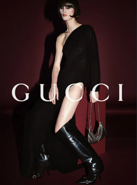 Fashion shoot: Woman in an elegant black one-shoulder dress and black boots, carrying a sparkling silver purse, against a dark red background.