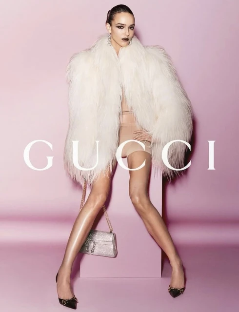 Fashion shot: A model in a cream fur coat poses against a pink backdrop, holding a glittering purse with a large logo. Gucci logo displayed.